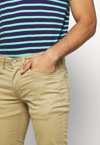 Khaki chinos with a slim fit, featuring side pockets, a button closure, and a smooth texture; paired with a navy and mint striped shirt.