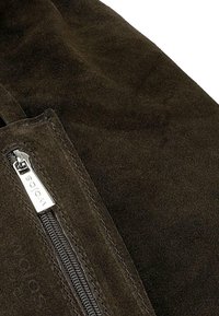Gros plan d’un tissu en daim marron foncé avec un curseur de fermeture éclair en métal gravé avec "Wojas" sur une poche cousue.
