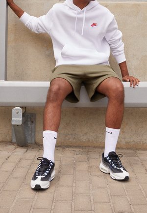 Persoon zit op een metalen bankje, draagt een witte Nike hoodie, olijfgroene korte broek, witte Nike sokken en zwart-witte sneakers met blauwe accenten.