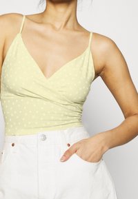 Woman wearing a pale yellow wrap top with small flower print and white high-waisted pants, with her right hand in a pocket.