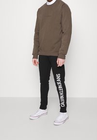 Hnědá mikina se vzorovaným logem, spojená s černými tepláky, které mají velký bílý nápis. Oblečeno s bílými teniskami. Minimalistický design.
