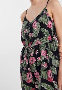 Mono floral con tirantes finos en negro, adornado con grandes flores de hibisco rosas y hojas verdes; cinturón en la cintura para ajuste.