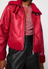 Veste rouge en similicuir avec un col et des poignets en sherpa, équipée de ferrures argentées, d'une fermeture éclair frontale et de sangles décoratives au niveau du cou.