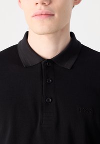 Μαύρο πόλο μπλουζάκι από υφασμάτινη υφή, με τριπλή κουμπί, δομημένο γιακά και διακριτό ανάγλυφο λογότυπο στο στήθος.