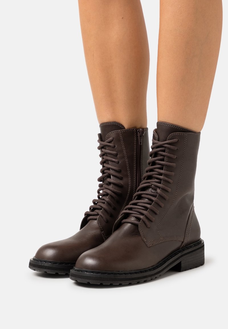 Paire de bottines en cuir marron foncé à lacets avec fermetures éclair latérales portées sur jambes nues sur fond blanc.