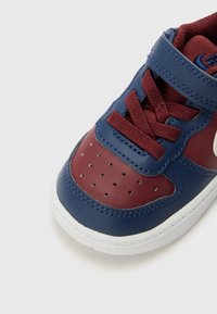 Sapatilhas de pele azul-marinho e bordô com colarinho almofadado, biqueira perfurada e sola branca. Apresentam uma fivela de velcro e fecho com atacadores.