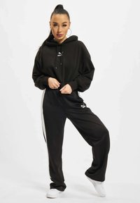 Sudadera negra con capucha y un logo, combinada con joggers negros que tienen franjas laterales blancas y zapatillas blancas. Tela suave y corte relajado.