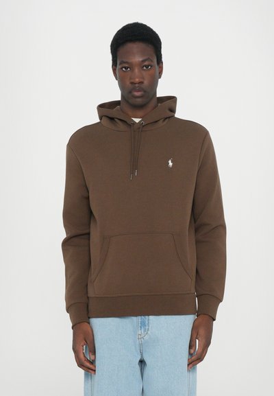 Bruine hoodie van zachte stof, met een kenguruzak, trekkoorden en een klein wit logo op de borst. Draagt met lichtblauwe jeans.