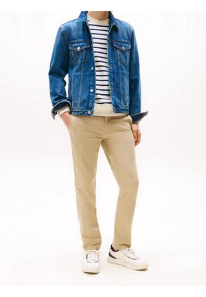 Mand iført blå denimjakke, hvid og marineblå stribet sweater, beige bukser og hvide sneakers, stående med hænderne i lommerne.