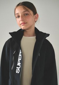 Svart zip-up jacka med hög krage och stor vit "SUPERDRY" logotyp. Har en enfärgad design, slät textur och sidofickor med dragkedja.