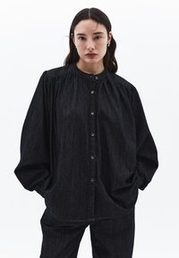 Černá džínsová košile s kulatým límcem, zapínáním na knoflíky a nařasenými rameny. Volný střih s širokými rukávy a jemnou strukturou.
