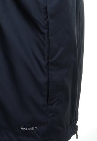 Marineblå Nike Shield-jakke i en glat, tekstureret kvalitet. Har en lynlås lomme og broderet logo nær kanten.