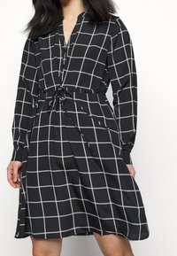 Černé šaty s bílým mřížkovaným vzorem, dlouhými rukávy, knoflíkovou légou, nastavitelným stahovacím pasem a volnou sukní. Hladká struktura látky.