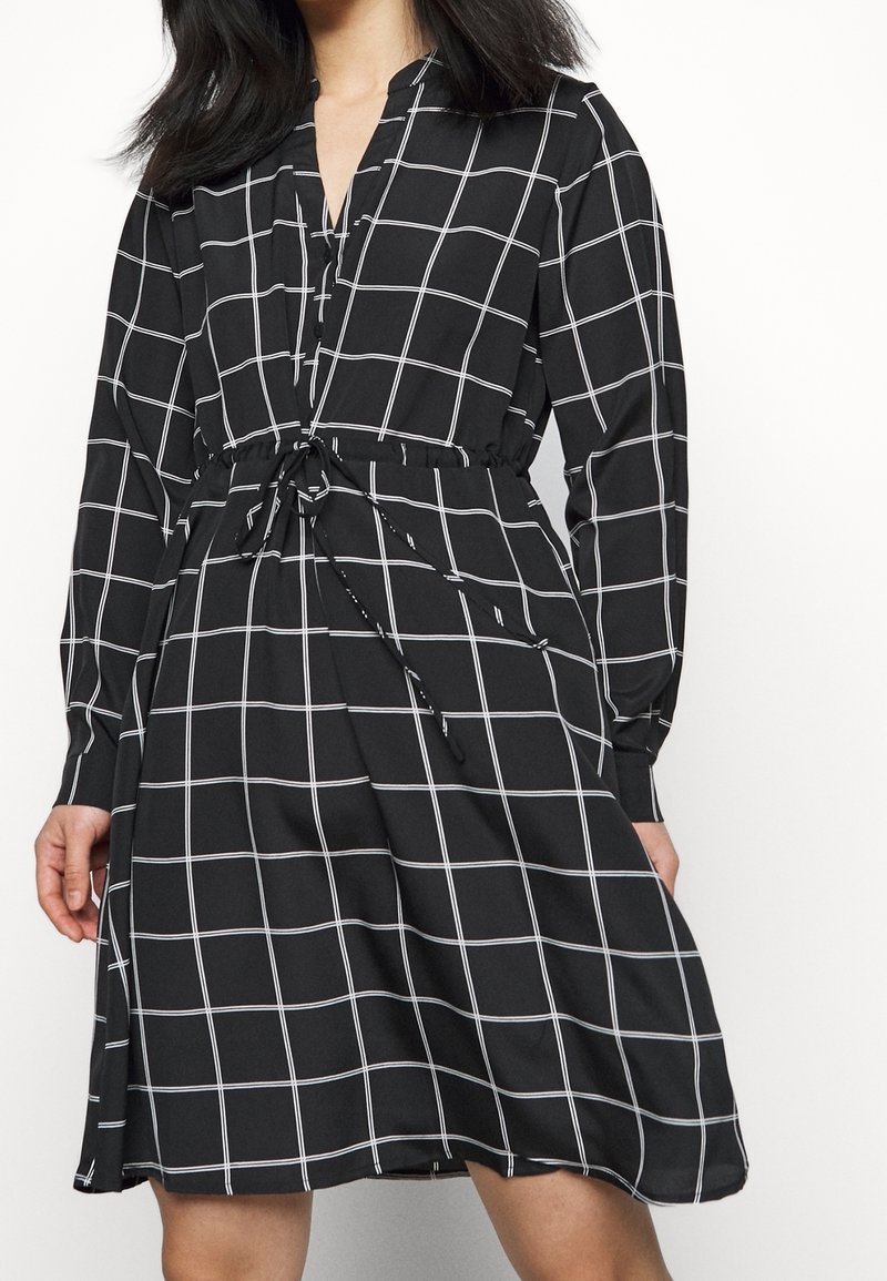 Černé šaty s bílým mřížkovaným vzorem, dlouhými rukávy, knoflíkovou légou, nastavitelným stahovacím pasem a volnou sukní. Hladká struktura látky.