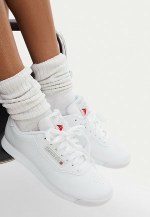 Piernas con zapatillas blancas Reebok y calcetines blancos arrugados, sentadas en una silla con los pies ligeramente levantados del suelo.