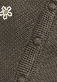 Strikket brunt stoff med ribbet kant, med en hvit brodert blomst og runde knapper langs siden for lukking.