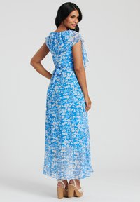 Kwiatowa maxi sukienka w kolorze niebieskim z białymi wzorami, z falbaniastego dekoltem, rękawami na kapelusz oraz wiązanym w pasie, uzupełniona o sandały na platformie.