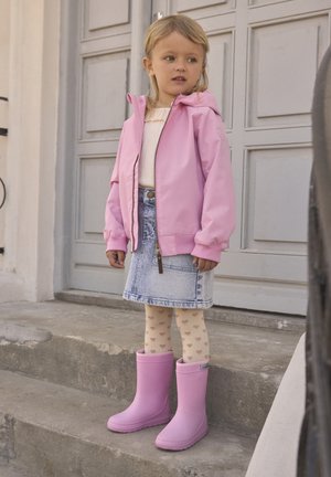 Ung pige iført en pink jakke, denim nederdel, strømpebukser med hjerte mønster og pink gummistøvler, der står på betontrin udenfor en grå dør.