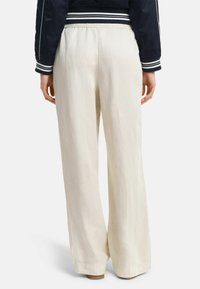 Weite Hosen aus cremfarbigem Stoff mit einer leichten, glatten Textur. Elastischer Bund und seitliche Taschen sichtbar; minimale Nähdetails.