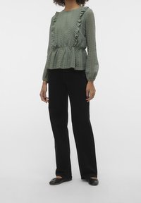 Blusa verde chiaro, strutturata, con dettagli a volant, maniche lunghe e vita arricciata; abbinata a pantaloni neri a gamba dritta e ballerine.
