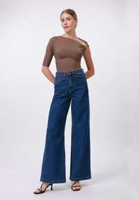 Rudas, prigludęs bodis su trumpomis rankovėmis ir šoniniu sagties akcentu; derinamas su aukštu liemeniu, plačiais mėlynais džinsais ir juodais aukštakulniais su dirželiais.