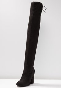 Botte noire en daim au-dessus du genou avec un talon épais, une texture lisse et un détail à lacets sur le dessus. Design sans coutures avec une silhouette ajustée.