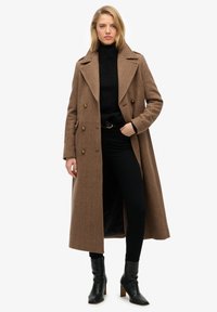 Brun frakke i fiskebenmønster med dobbeltradede knapper, spidse revers og sidelommer, kombineret med en sort rullekrave og sorte bukser med slank pasform.