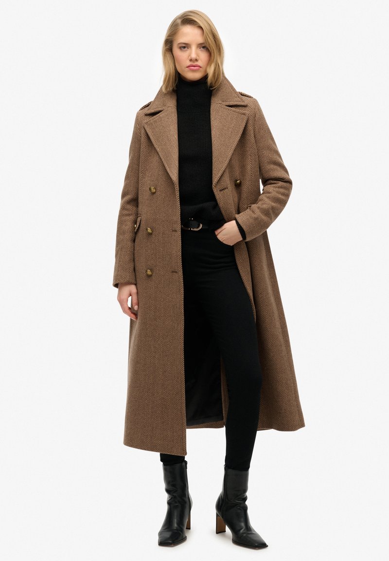 Brun frakke i fiskebenmønster med dobbeltradede knapper, spidse revers og sidelommer, kombineret med en sort rullekrave og sorte bukser med slank pasform.