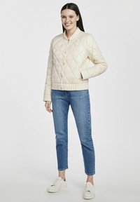 Pikowana kremowa kurtka z kołnierzykiem, z przodu z zapięciem na napy i bocznymi kieszeniami, zestawiona z niebieskimi dżinsami i białymi sneakersami.