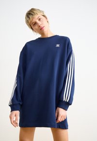 Marinblå oversized sweatshirtklänning i bomull, med rundad halsringning, långa ärmar med vita ränder och en liten logotyp på bröstet.