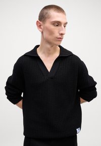 Schwarzer Strickpullover mit V-Ausschnitt und geripptem Kragen, mit lockeren Ärmeln und strukturiertem Material. Kleines Logotag am Saum.