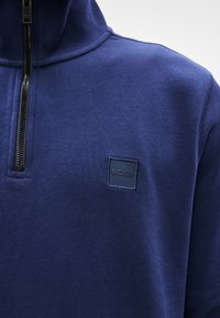 Πουλόβερ σε ναυτικό μπλε χρώμα με υψηλό γιακά και κλείσιμο με μισό φερμουάρ. Διαθέτει υφασμάτινο τετράγωνο πατς με το λογότυπο "BOSS" στο στήθος. Μαλακό ύφασμα.