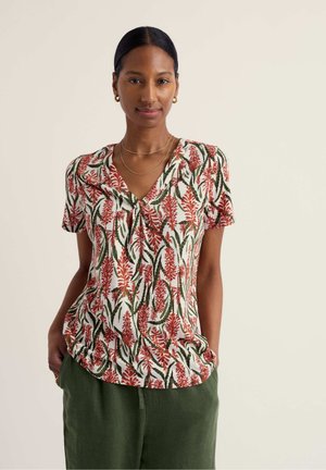 Femme aux cheveux foncés portant un haut à manches courtes à motifs floraux avec des fleurs rouges et des feuilles vertes, vêtue d'un pantalon vert, debout les mains dans les poches.