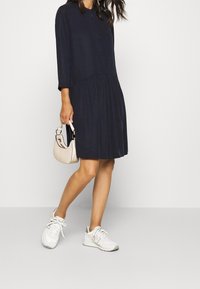 Donna che indossa un abito blu navy con maniche lunghe e bottoni, sneakers bianche e tiene in mano una piccola borsa beige mentre cammina su uno sfondo chiaro.