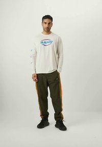 Mand står med hænderne i lommene iført en hvid langærmet skjorte med "ARMY" logo og mørke bukser med orange sidelister og sorte sneakers.