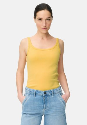 Kobieta o ciemnych włosach, ubrana w żółty top na ramiączkach i jasnoniebieskie dżinsy, stoi z rękami w kieszeniach, zwrócona przodem na jednolitym tle.