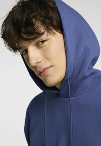 Felpa con cappuccio blu navy dotata di lacci, morbida al tatto e dalla vestibilità rilassata, con un ampio cappuccio e cuciture minime, indossata da una persona.