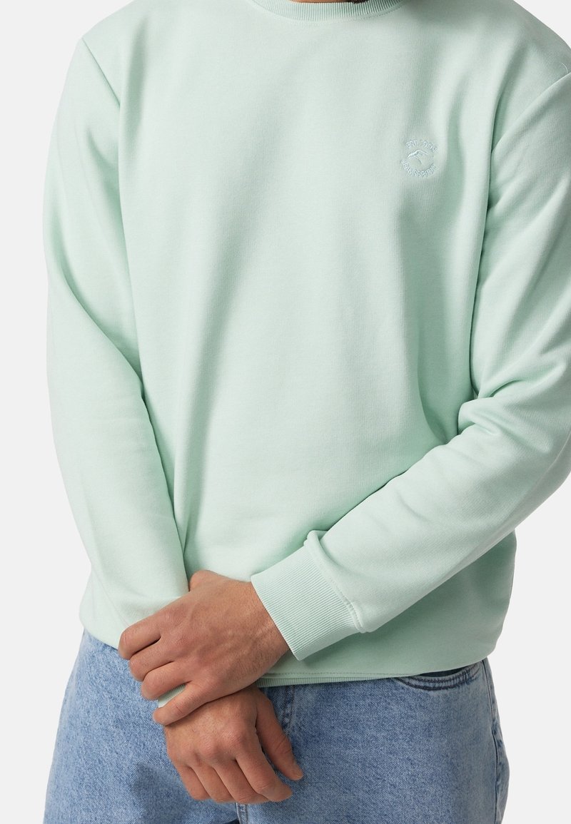 Homme portant un sweat-shirt vert clair avec des poignets côtelés et un petit logo brodé, associé à un jean bleu clair, les mains jointes devant.
