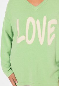 Maglione oversize verde con scollatura a V, caratterizzato da una grafica "LOVE" color crema in un font giocoso. Tessuto morbido e vestibilità rilassata.