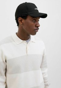Weiß gestreiftes Poloshirt mit strukturiertem Finish, ausgestattet mit einem Knopfkragen. Getragen mit einer schwarzen Baseballmütze, die "JJ Studio" in Weiß zeigt.