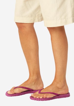 Ben klædt i beige shorts og lilla flettede flip-flops med strukturerede såler på en neutral baggrund.