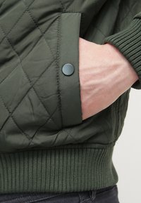 Jachetă verde matlasată cu manșete striate, având un buzunar lateral asigurat cu un nasture negru tip snap. Textura pare netedă, materialul este ușor.