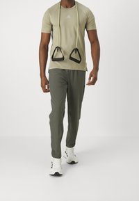 Hombre con camiseta beige de Adidas y pantalones grises de Adidas sosteniendo mangos de banda de resistencia, caminando hacia adelante sobre un fondo blanco liso.