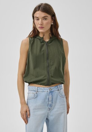 Kvinde iført ærmeløs olivengrøn lynlås-hoodie og lyseblå højttaljede jeans, stående mod en ensfarvet baggrund.