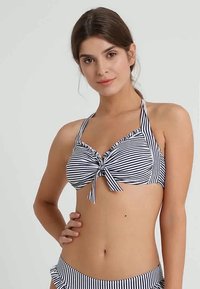 Jovem mulher com um top de bikini halter listrado em preto e branco, com um nó na frente e calções combinando, sorrindo levemente, com a mão na anca.
