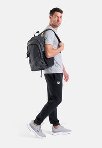 Zaino nero e grigio con logo, spallacci imbottiti, forma strutturata e tasche con cerniera. Indossato da un modello maschile in piedi con abbigliamento casual.