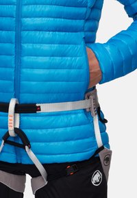 Blå vadderad jacka med horisontell sömnad, med dragkedjefickor och bärs över en klättringssele. Materialet ser blankt och lättviktigt ut.