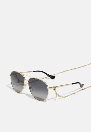 Gafas de sol aviador doradas con lentes de gradiente oscuro, que cuentan con una cadena decorativa y una punta de goma negra en las varillas. Nombre de la marca en las monturas.