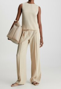 Beige ärmlös topp i kombination med vida byxor, båda gjorda av ett mjukt tyg. En matchande beige handväska hålls i ena handen.