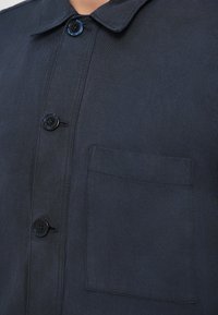 Tengerészkék, galléros inget, négy gombbal és egy zsebfedéllel. Tartós, textúrált anyagból készült.